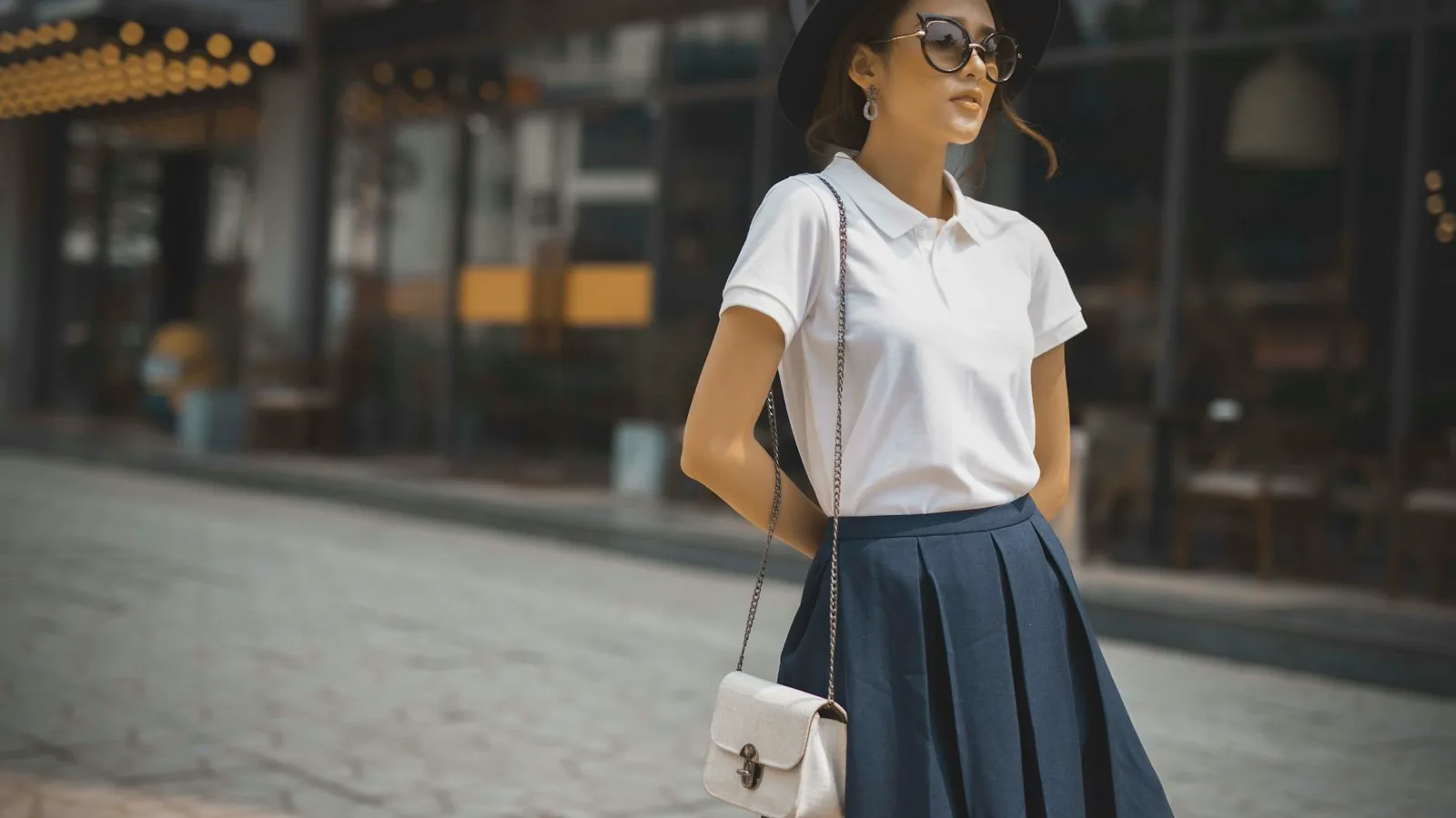 Stylish woman in white collared polo shirt, navy pleated skirt, sun hat, sunglasses, and crossbody bag demonstrating smart casual golf attire
